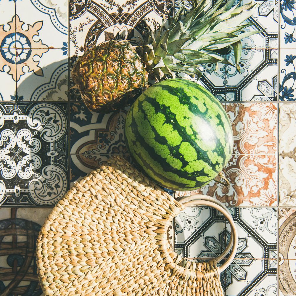 Thatched bag laying on pattered tiles with watermelon and pineapple, square crop