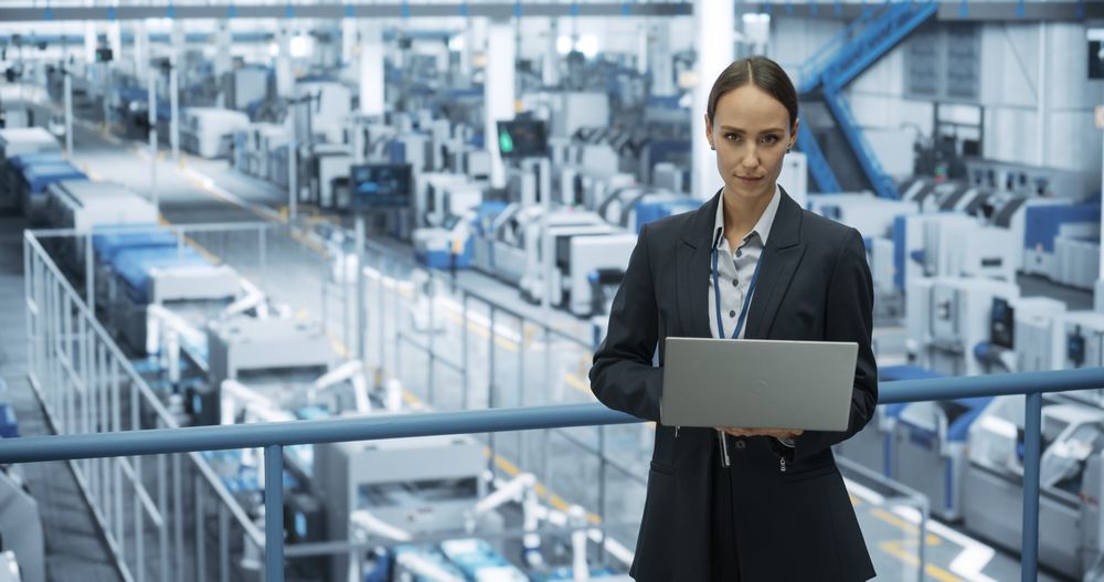 Young Female Engineer Standing on a Platform, Using Laptop Computer at an Electronics Factory. Machines are Undergoing Maintenance, Technician Monitoring Updates Through Online Software