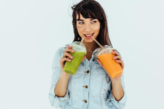 Portrait of beautiful woman drinking fresh orange and green juices on light background