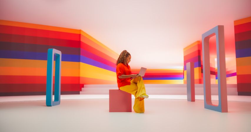 Black Woman in Bright Outfit Using Laptop Computer While Sitting in Geometric Abstract Environment. Creative Young Female Joyfully Using the Internet in a Studio with Colorful Background