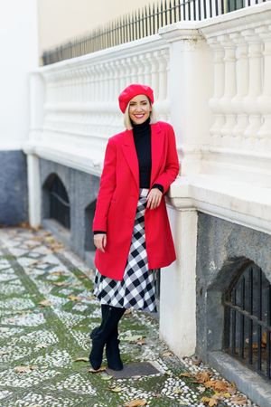 Full body of smiling female in trendy red coat and beret looking at camera standing in city