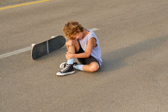 Cheerful skater sitting near skateboard on road