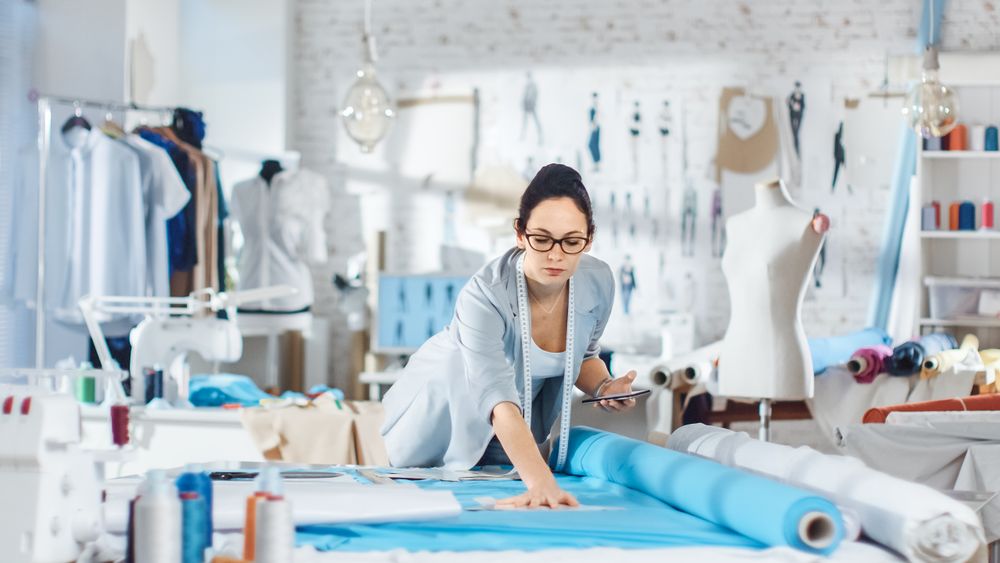 Brunette fashion, designer,  Lays Out Templates on the fabrics and Consults Her Smartphone.