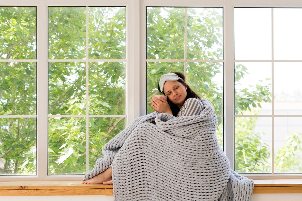 Woman enjoys tranquility, wrapped in gray blanket by window.