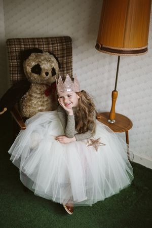 Young girl in light colored dress sitting on chair beside stuffed toy