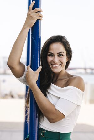 smiling young woman hanging on a pole in the street while looking camera