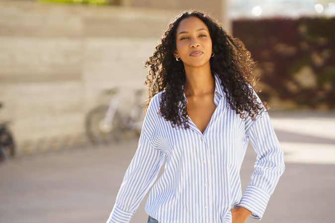 A confident young woman dressed in a stylish striped shirt is strolling outdoors blissfully