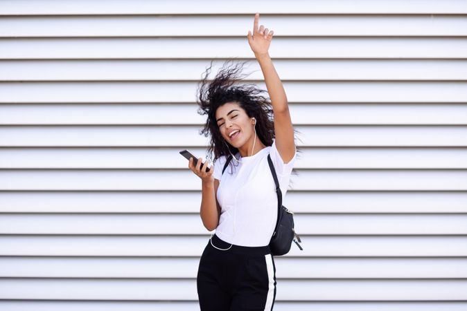 Happy Arab woman in sport clothes with curly hair standing in front of wall dancing and pointing up