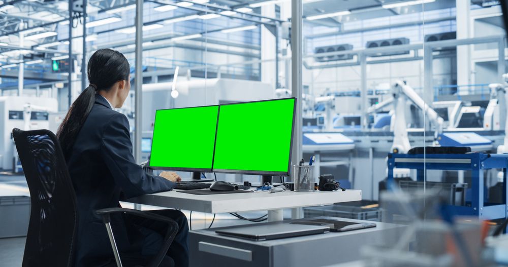 Female Robotics Technician Working on a Desktop Computer with Green Screen Chromakey Mock Up Display in a Factory Office. Technological Research and Development Facility with Autonomous Assembly Line
