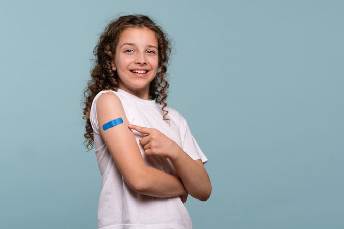 Smiling girl pointing at blue bandage after receiving vaccine.