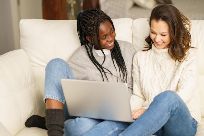 Two women in jeans and sweater laughing and watching something together on a sofa on a pc