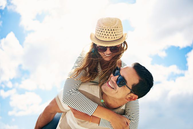 Woman piggy backing on boyfriend’s back against a blue sky with clouds