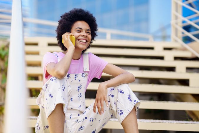Smiling woman in floral overalls sitting on stairs taking phone call