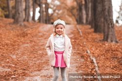 Young girl wearing pink jacket and gray hat standing on brown tree leaves 0LWODb