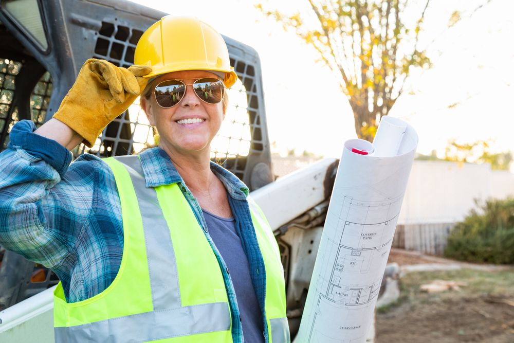 Smiling Female Worker Holding Technical Blueprints Near Small Bulldozer At Construction Site