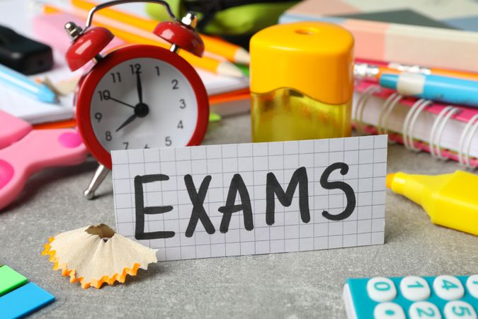 The word “Exams” surrounded by clock and stationary on desk