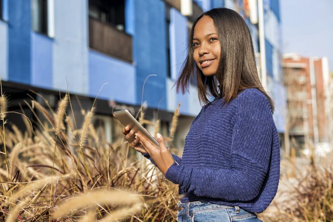 Smiling female in sweater standing outside blue building in long grass with tablet