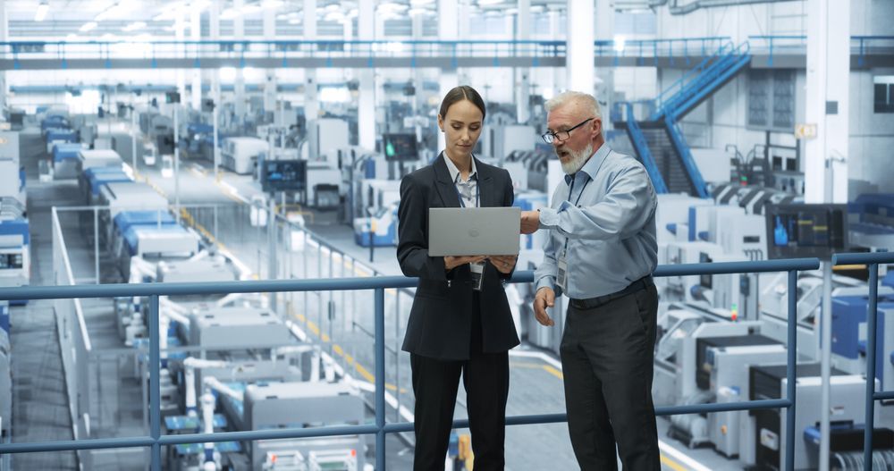 Male Engineer and a Female Technician Having a Conversation and Using Laptop Computer. Specialists Monitoring, Analyzing Conditions at a Modern Electronics Factory with Automated Robotic Arms