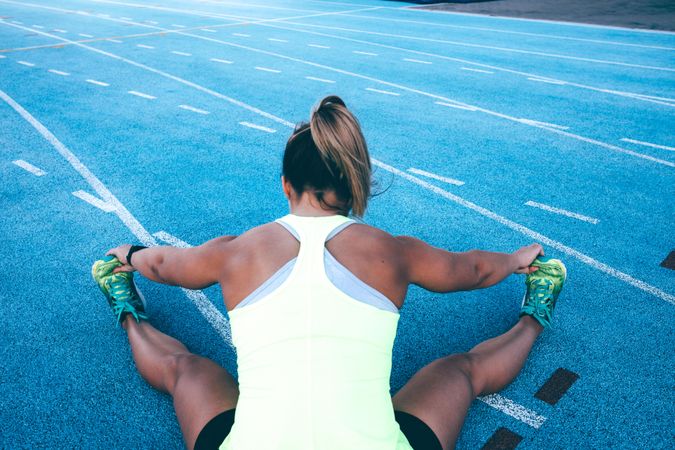 Young woman sitting on the lands of a track field stretching her legs