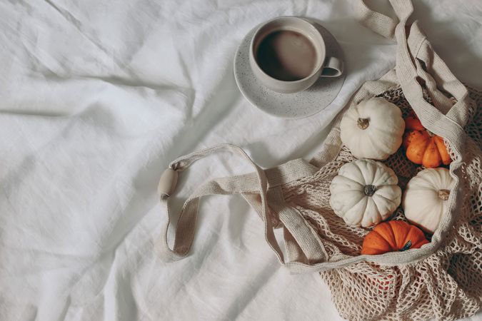 Little pumpkins, reusable cotton mesh bag, cup of coffee on linen fabric table, flat lay, top view