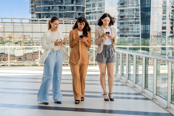 Three businesswomen are walking while using smartphones outdoors. All women are dressed in business attire..