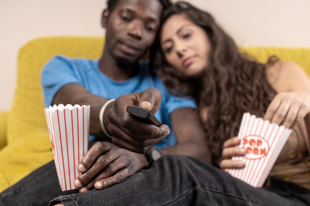 A tired and bored couple scrolling endlessly with a remote control, struggling to find a movie to watch at home.