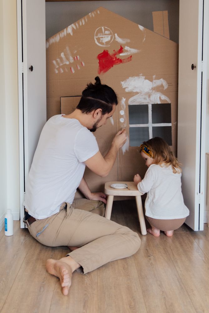 Back view of father and young daughter building a small house out of cardboard at home