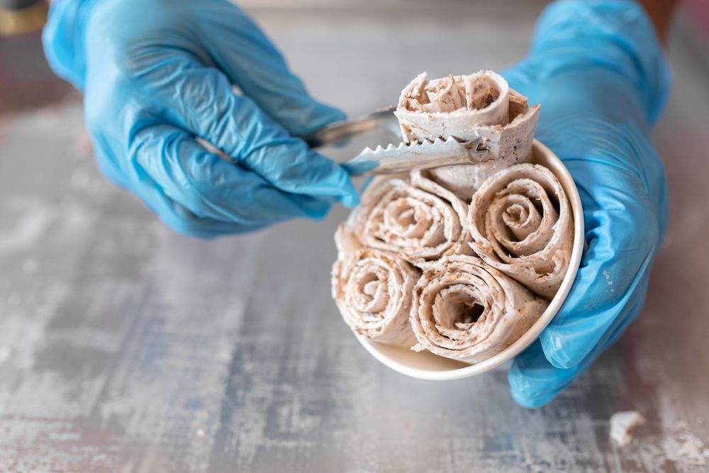 A worker is putting chocolate ice cream rolls in a tub using tongs and latex gloves