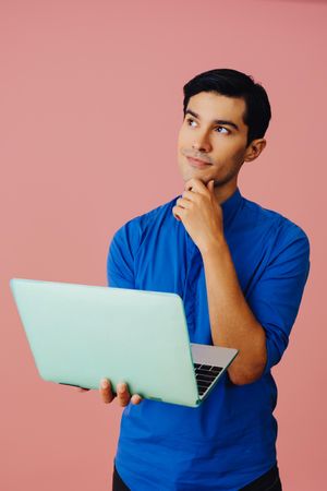 Vertical shot of contemplative Latino man looking up while holding laptop in pink room, copy space