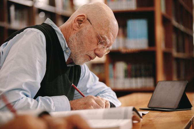 Older man writing in his book sitting in a library