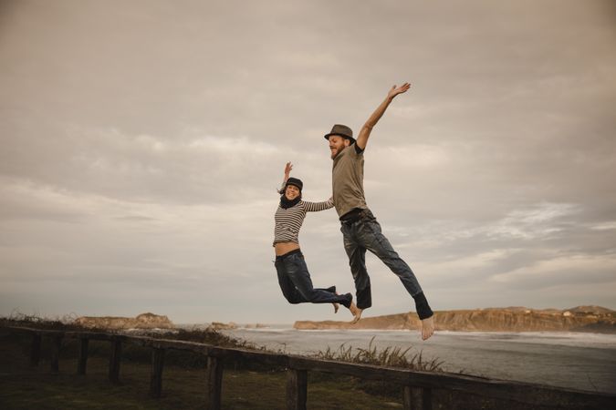 Couple holding hands and jumping on bench on shore near water