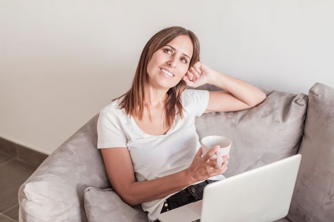 Woman thinking on sofa with a cup of coffee working on her laptop in the morning