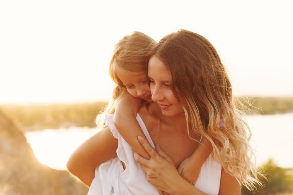 Portrait of young girl on woman’s back in front of river at dusk