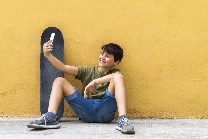 Boy sitting on ground leaning on a yellow wall and taking a selfie