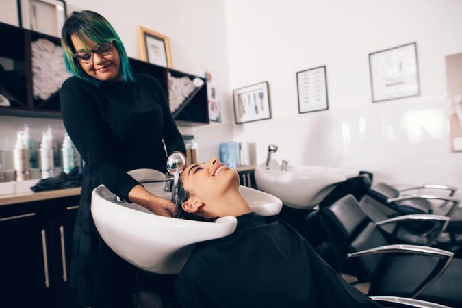 Young hairdresser with bright hair rinsing client’s hair in salon sink