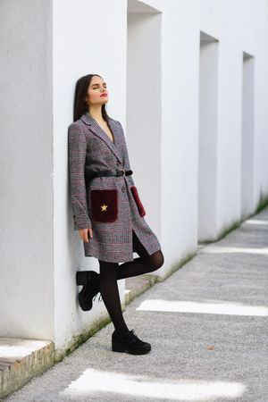 Female leaning on bright wall with eyes closed outside in belted winter coat dress