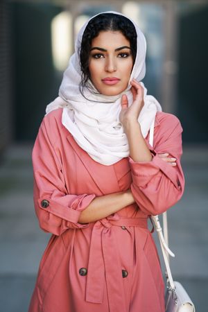 Female in headscarf and pink trench coat standing confidently with with one hand to her face