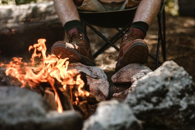 Older man in boots warms his feet by the fire at camping site