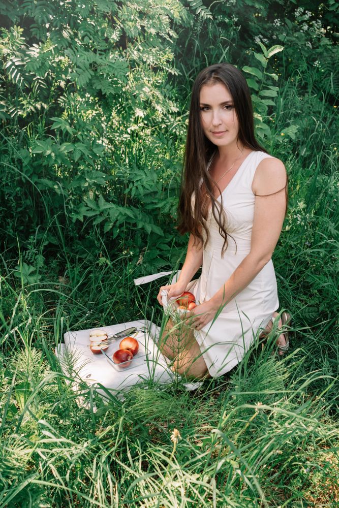 Woman in light dress kneeling on grass beside textile with fruit slices