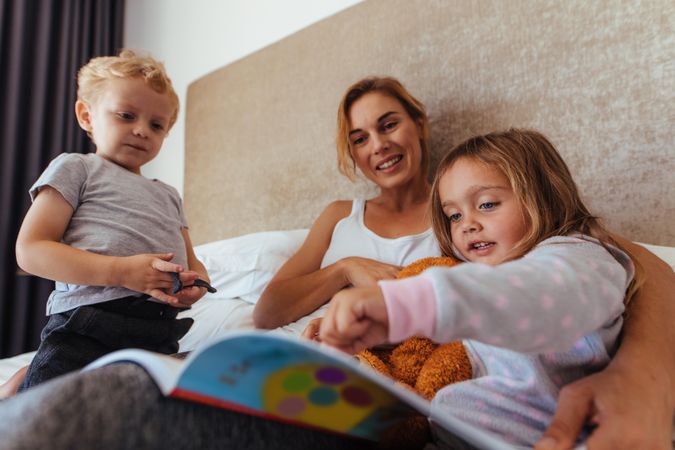 Cute little girl pointing at storybook while sitting with her mother and brother on bed
