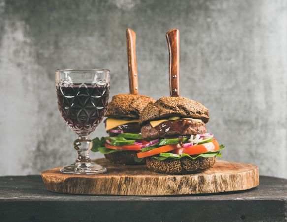 Two cheeseburgers skewered with knives, stacked with fresh vegetables, on wooden board, with wine