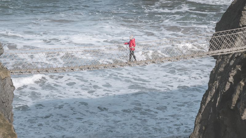 Tourist walking on rope bridge suspended between cliffs in Northern Ireland