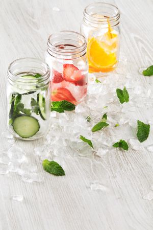 Three mason jars of infused water with distilled water bottle surrounded with ice and mint leaves