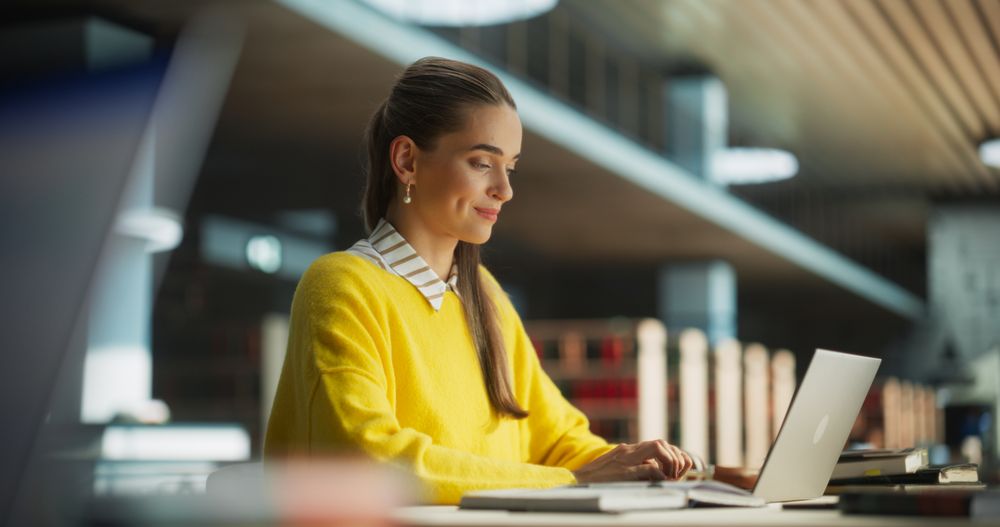 Female Student in Yellow Jumper Working on Her School Thesis on a Laptop Computer. Young Smart Woman Studying Online in a Quiet Public Library, Writing Down Report in Notebook