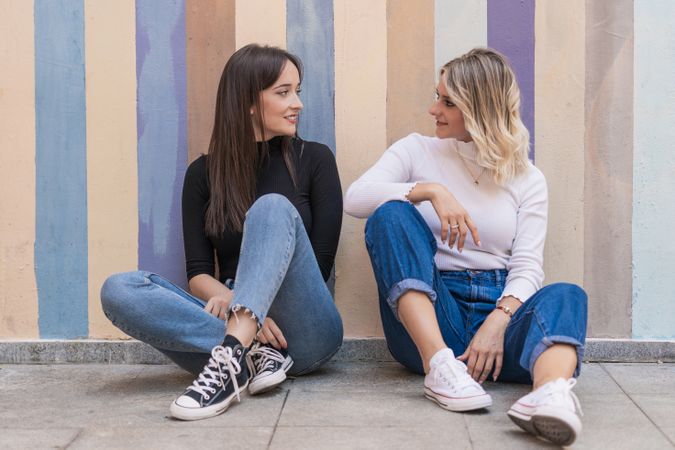 Happy girlfriends sitting against painted urban wall