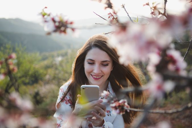 Young happy woman taking selfie on smartphone near trees on hill