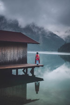 Person standing beside house on dock over lake under cloudy sky