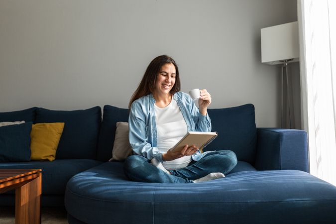 Cheerful pregnant woman drinking beverage on sofa at home
