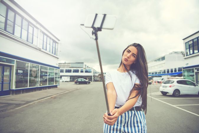 Woman with long brown hair posing in street with selfie stick for photo