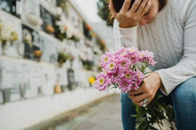 Grieving woman touching forehead while sitting near columbarium wall in cemetery